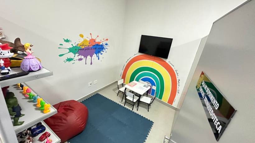 Interior de uma brinquedoteca infantil, com paredes decoradas em cores vibrantes, uma mesa e cadeiras para crianças, e prateleiras com brinquedos e pelúcias.