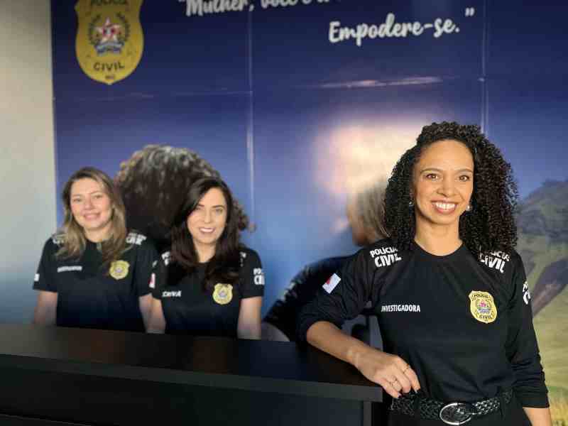 Três mulheres policiais civis, uma delas em primeiro plano sorrindo, em frente a um mural da Polícia Civil com lema inspirador sobre empoderamento feminino.