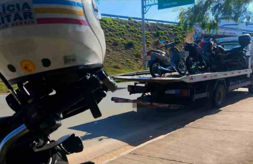 Imagem de uma motocicleta da Polícia Militar em primeiro plano, com um caminhão reboque ao fundo transportando várias motocicletas apreendidas em uma estrada.