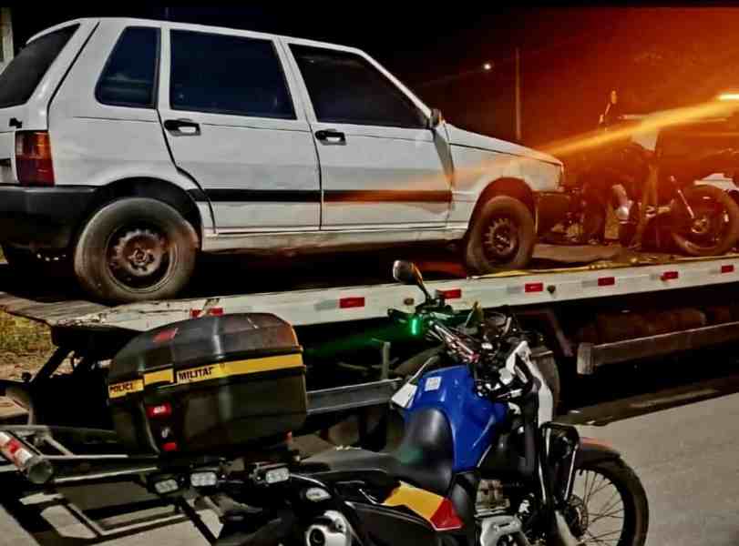Uma caminhonete branca sendo rebocada com uma motocicleta ao lado, em um ambiente noturno.