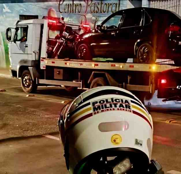 Imagem de uma motocicleta sendo carregada em um caminhão de reboque, com um carro em cima da plataforma. Ao primeiro plano, há um capacete da Polícia Militar de Minas Gerais.