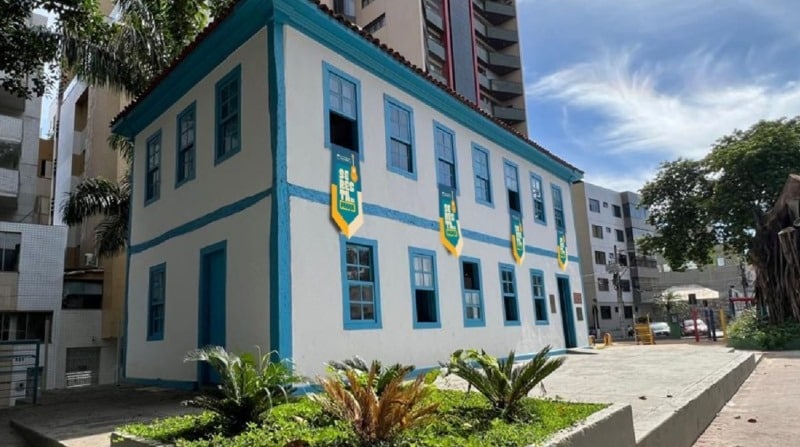 Fachada de um edifício histórico com pintura branca e detalhes em azul, cercado por plantas e em meio a prédios modernos.