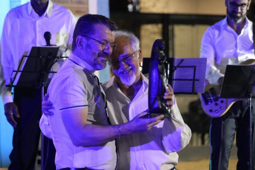 Dois homens sorrindo e se abraçando em um palco, segurando um objeto, com músicos ao fundo.