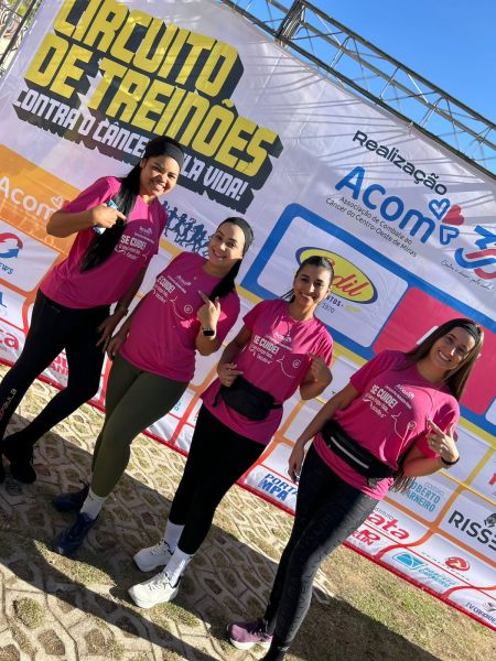 Quatro mulheres sorrindo, vestindo camisetas rosas, posando para a foto em um evento de conscientização contra o câncer de mama, com um banner ao fundo.