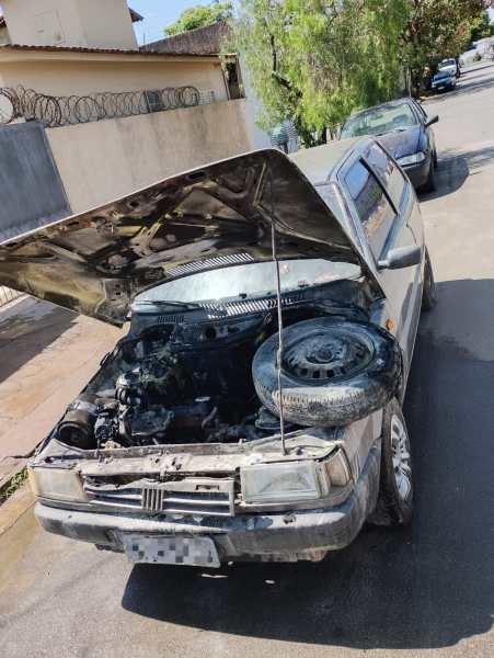 Carro com o capô levantado e motor exposto em uma rua, possivelmente após um incêndio.