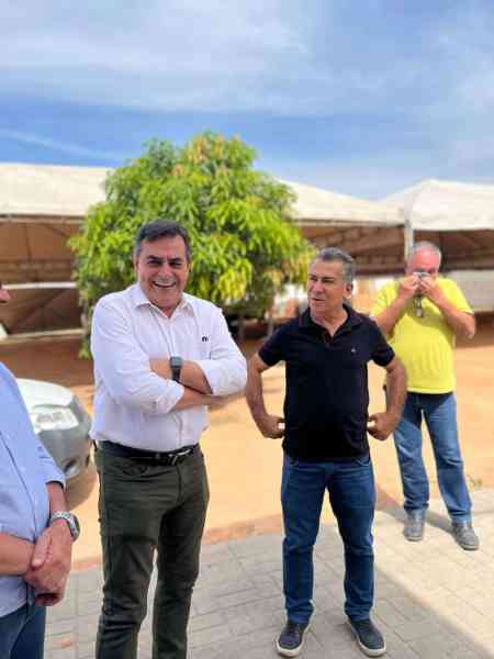 Dois homens sorridentes em um evento ao ar livre em Nova Serrana, com um fundo de árvores e tendas. Um homem está de braços cruzados, enquanto o outro está ao seu lado, ambos conversando. Um terceiro homem aparece ao fundo, tirando fotos.