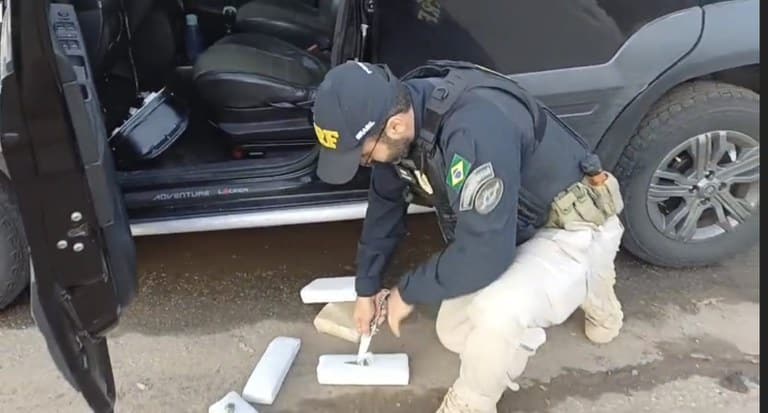 Policial em uniforme da PRF inspecionando substâncias apreendidas ao lado de um veículo Fiat Idea Adventure, em um local de estrada.