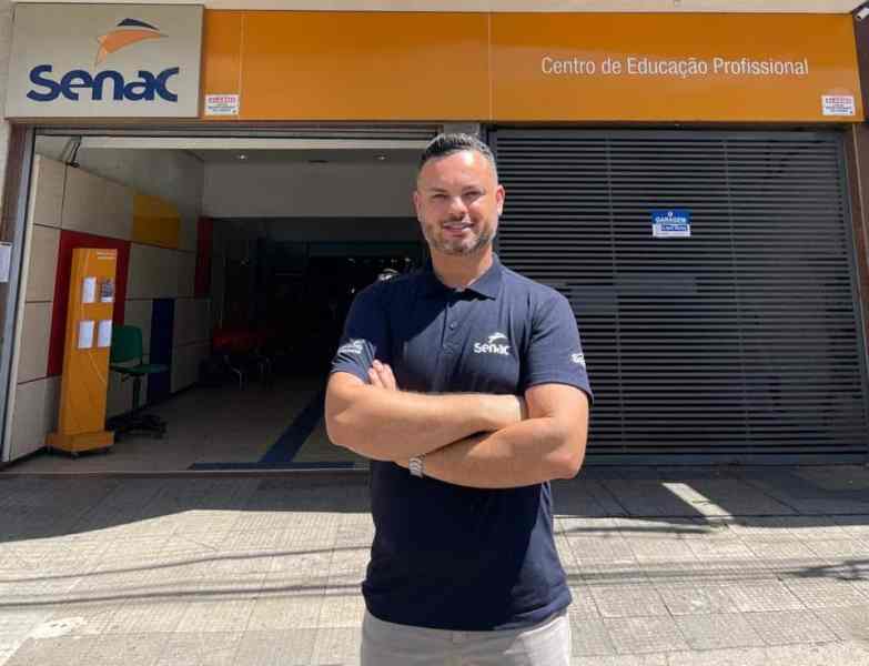 Homem sorridente em frente ao prédio do Senac, que possui placa com o nome 'Centro de Educação Profissional'.