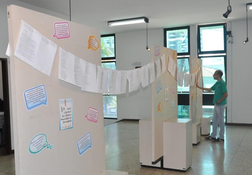Sala expositiva da Escola do Legislativo Dr. Deusdedith Afonso Carrilho, com painéis expositores e textos pendurados, e um participante interagindo com o conteúdo.