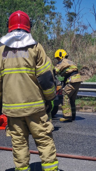Dois bombeiros em uniformes de proteção, um com capacete amarelo e outro com capacete vermelho, estão em uma rodovia durante simulado de resgate após um acidente.