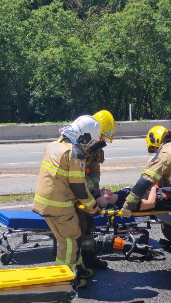 Bombeiros militares atendendo uma vítima simulada em uma estrada durante um exercício de resgate relacionado a acidentes com produtos perigosos.