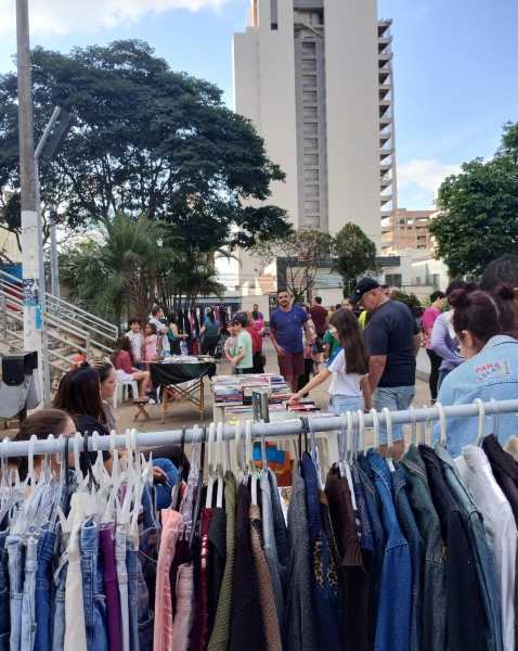 Vista da Praça do Santuário durante a Feira Astral, com pessoas interagindo e um varal de roupas expostas ao primeiro plano, em um ambiente vibrante e acolhedor.