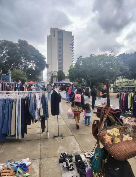 Visão geral da Praça do Santuário durante a Feira Astral, com barracas de roupas e artesanato à mostra, pessoas caminhando e um edifício ao fundo.