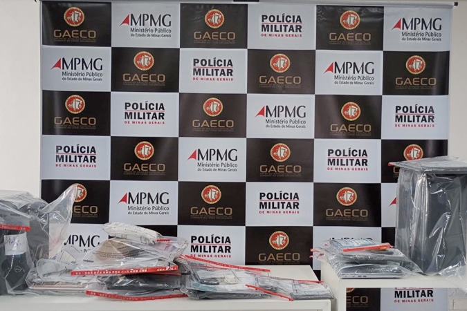 Mesa com pacotes plásticos contendo documentos e materiais de prova em frente a um fundo com logos do MPMG, GAeco e Polícia Militar.