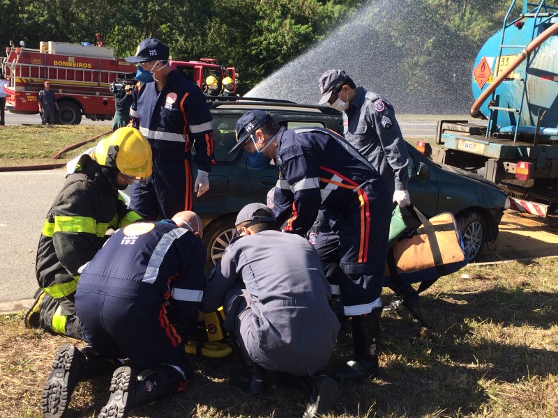 Simulado de acidente com múltiplas vítimas, com equipes de resgate, incluindo bombeiros e agentes de saúde, atendendo a uma situação de emergência ao lado de um veículo.