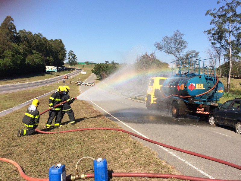 Cena de simulado de acidente com produtos perigosos, com bombeiros usando mangueiras para combater um possível incêndio em um caminhão tanque na rodovia MG-050. A equipe de emergência está em ação, criando um arco-íris a partir da água.