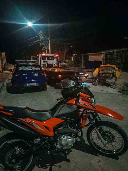 Motocicleta laranja estacionada em uma rua à noite, com um veículo de guincho ao fundo e iluminação pública.