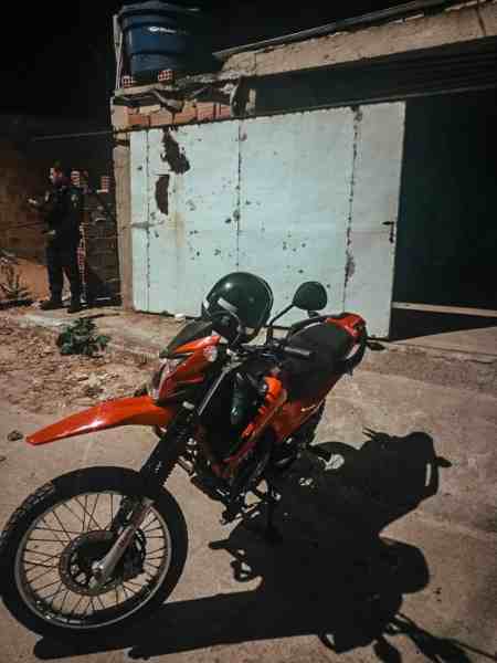 Motocicleta vermelha estacionada em frente a um galpão com portas azuis, enquanto policiais realizam a abordagem ao fundo.