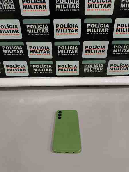 Imagem de um celular verde sobre uma mesa com fundo de placas da Polícia Militar de Minas Gerais.
