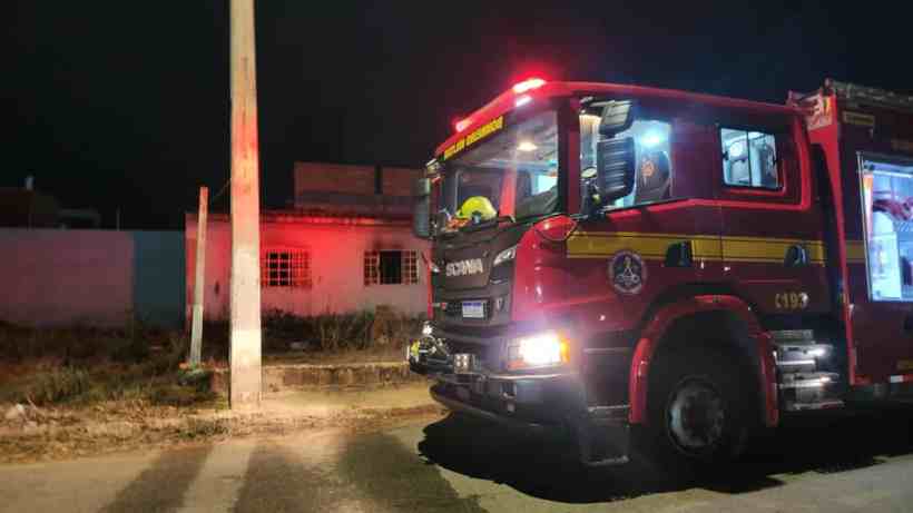 Caminhão de bombeiros estacionado em uma rua à noite, com luzes vermelhas e amarelas acesas, próximo a uma residência aparentemente abandonada.