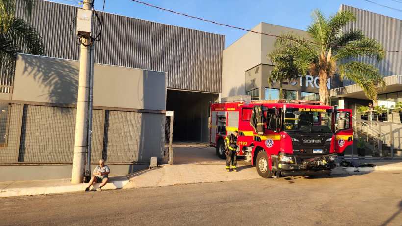 Um caminhão de bombeiros estacionado em frente a uma fábrica com paredes metálicas e uma entrada ampla. Um bombeiro está próximo ao veículo, enquanto um homem observa sentado na calçada.