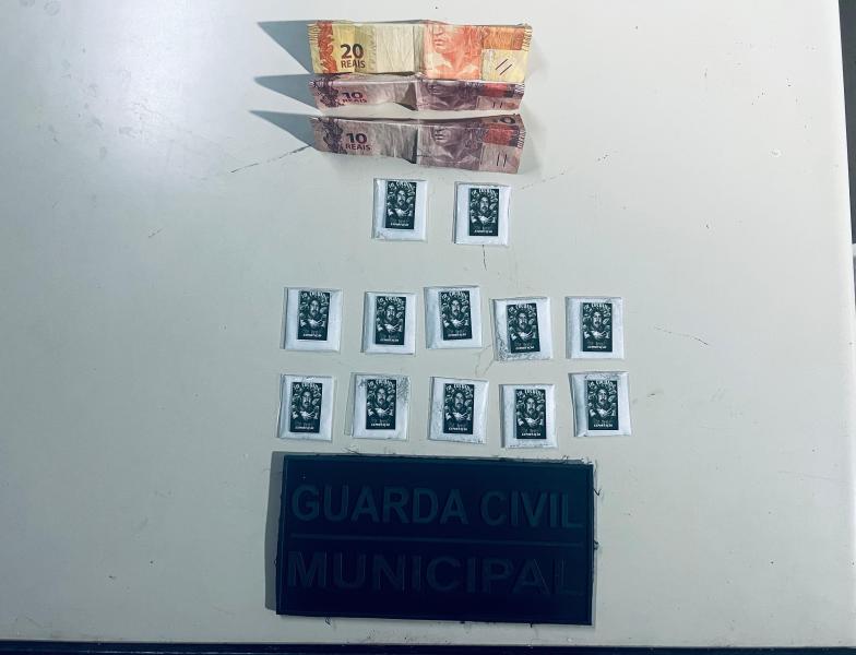 Notas de dinheiro brasileiro (duas de 20 reais e duas de 10 reais) dispostas acima de embalagens de substâncias, juntamente com uma placa da Guarda Civil Municipal.