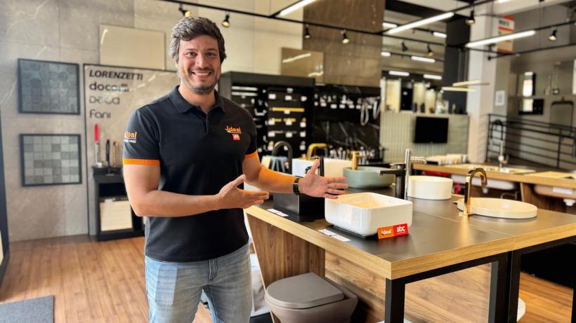 Homem sorridente apresentando diferentes pias e torneiras em uma loja de materiais de construção, com produtos da marca Ideal e ABC em destaque ao fundo.