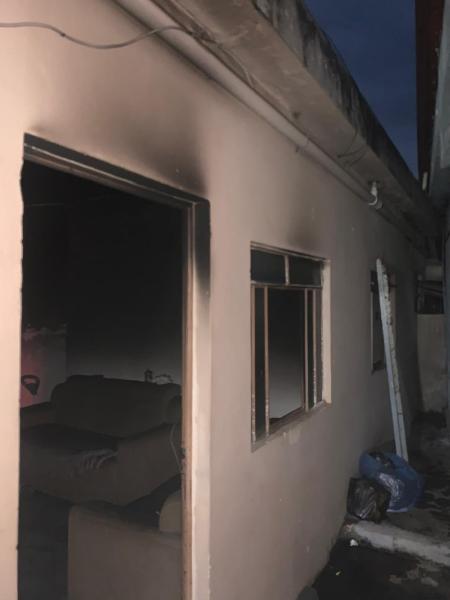 Parte externa de uma residência com marcas de fogo, incluindo paredes queimadas e janelas danificadas. O ambiente aparenta ter sofrido um incêndio, com móveis visíveis ao fundo.