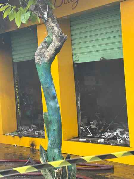 Uma loja de cosméticos em Divinópolis apresenta danos significativos após um incêndio, com fumaça visível e prateleiras destruídas, enquanto uma árvore próxima é parcialmente coberta por cordas de isolamento.