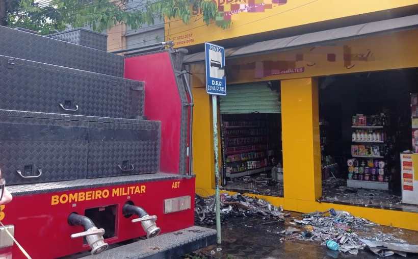 Caminhão do Corpo de Bombeiros estacionado em frente a uma loja de cosméticos danificada por incêndio, com a fachada amarela e fumaça visível.