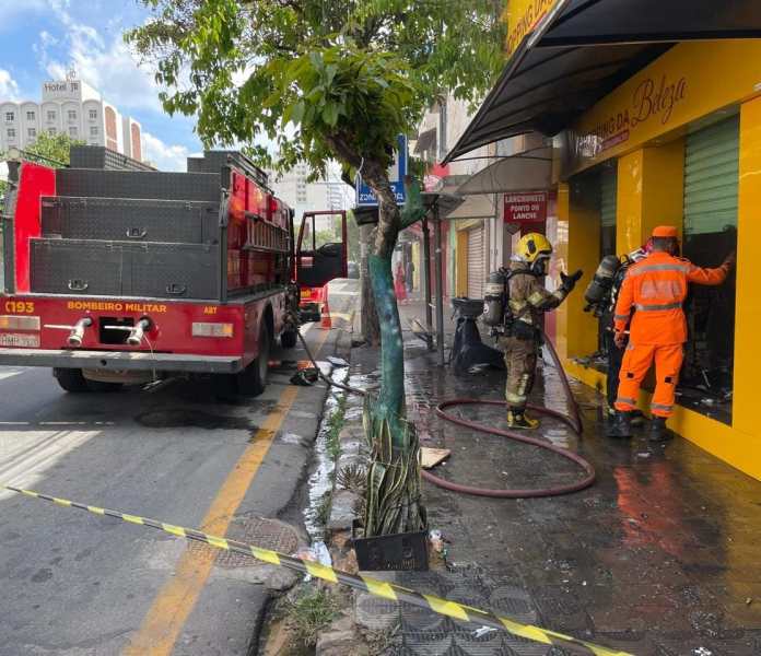 Caminhão dos bombeiros estacionado na rua com bombeiros em ação próximo a uma loja de cosméticos, após um incêndio.
