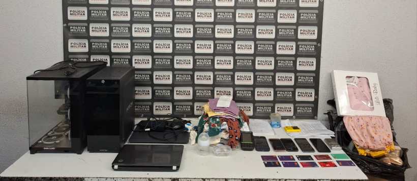 Mesa com diversos materiais apreendidos pela Polícia Militar, incluindo computadores, celulares, cartões e mercadorias, em fundo com logotipo da Polícia Militar.