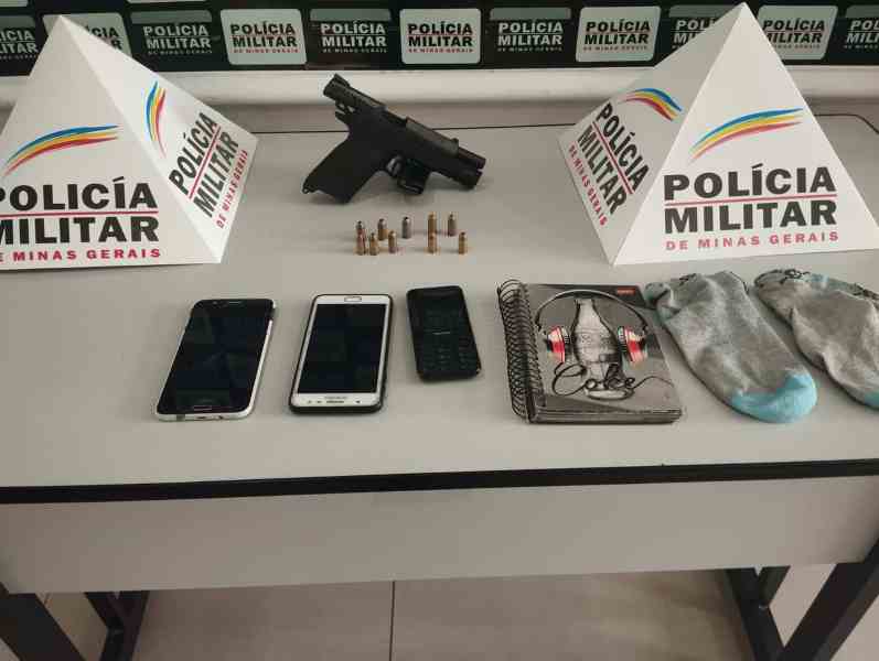 Mesa com materiais apreendidos pela Polícia Militar, incluindo uma pistola, munições, celulares, caderno e meias, com placas da Polícia Militar de Minas Gerais ao fundo.