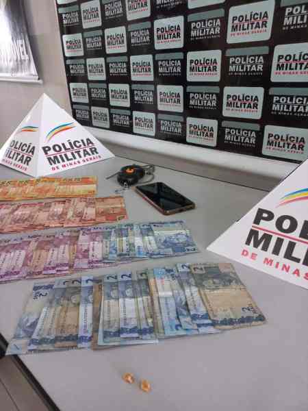 Dinheiro apreendido durante a 'Operação Incrédulos', com notas de várias denominações, um celular e os triângulos de sinalização da Polícia Militar de Minas Gerais ao fundo.