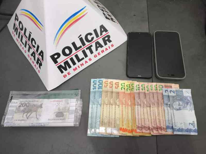 Notas de diversos valores, incluindo uma cédula de R$200,00, em frente a um material com a identificação da Polícia Militar de Minas Gerais, ao lado de dois celulares.
