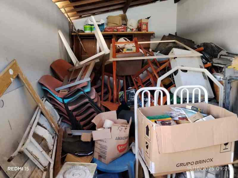 Ambiente desorganizado com cadeiras empilhadas, caixa de papelão e objetos acumulados, retratando condições inadequadas de moradia.