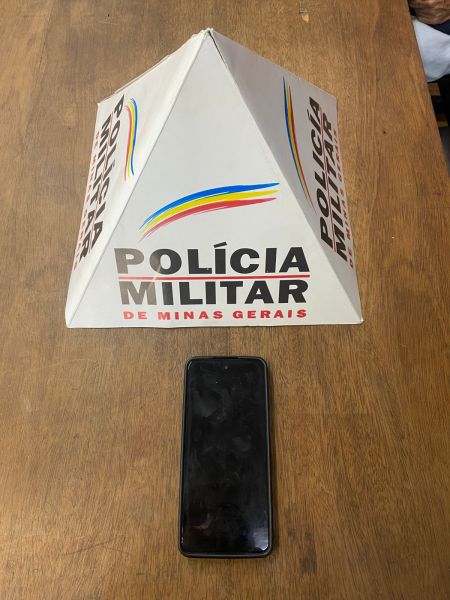 Um celular Motorola Moto G53 em cima de uma mesa com uma estrutura piramidal da Polícia Militar de Minas Gerais ao fundo.