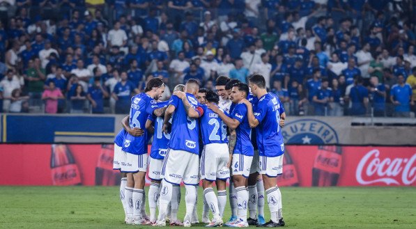 Jogadores do Cruzeiro em um momento de união durante o jogo, com camisas azuis e brancas, em um estádio cheio de torcedores ao fundo.