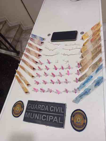 Mesa com dinheiro em espécie e porções de drogas, incluindo crack e cocaína, ao lado de um celular e placas da Guarda Civil Municipal.