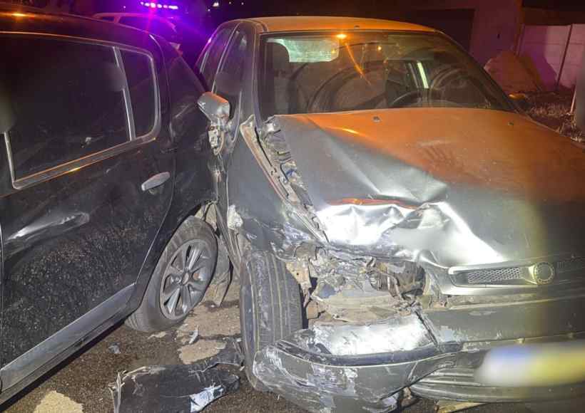 Imagem de um acidente de trânsito envolvendo um Fiat Palio e um Renault Sandero. O Palio apresenta danos significativos na parte frontal após colidir com o Sandero, que está estacionado. Luzes de emergência são visíveis ao fundo.