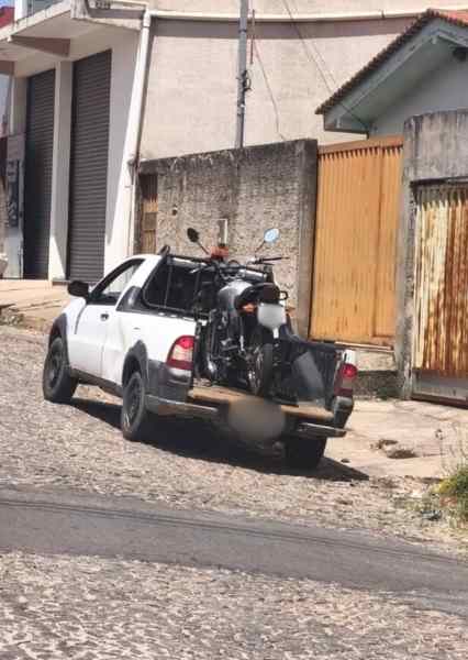 Uma caminhonete branca estacionada em uma rua de paralelepípedos, com uma motocicleta carregada na caçamba.