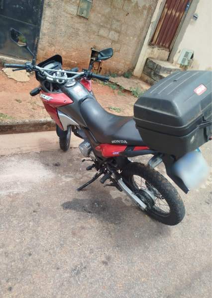 Uma motocicleta Honda na cor vermelha, estacionada em uma rua, com um baú na parte de trás.