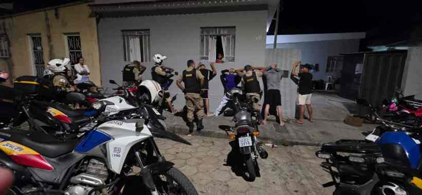 Agentes da Polícia Militar em operação noturna, revistando indivíduos em uma área urbana, com várias motocicletas estacionadas ao redor.