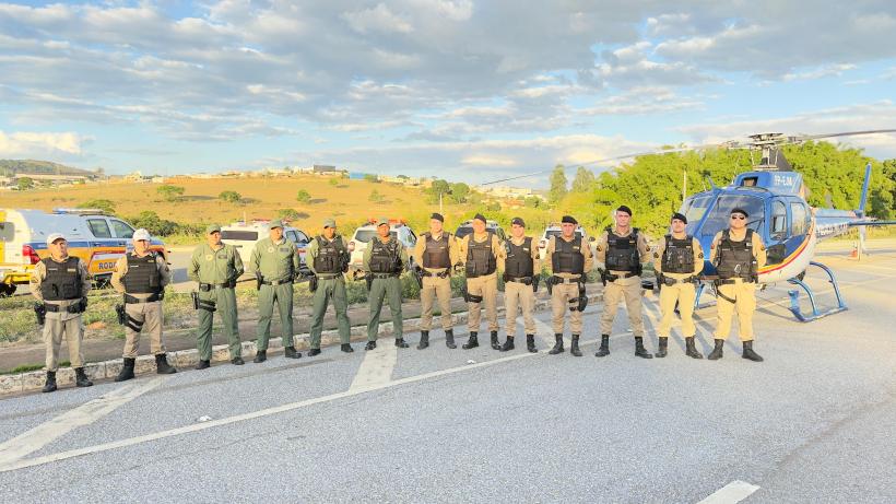 Policiais militares em uniforme tático alinhados em uma estrada, com um helicóptero da Polícia Militar ao fundo e veículos de serviço na lateral.