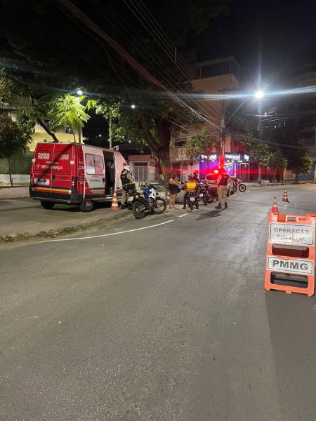 Policiais militares realizam abordagem em motocicletas durante operação noturna em Divinópolis, com viatura e sinalização policial visíveis.