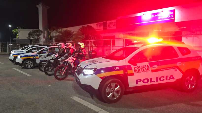 Viatura da Polícia Militar de Minas Gerais estacionada em frente ao sistema de segurança, com luzes de emergência acesas. Motocicletas policiais estão posicionadas ao lado dos carros.