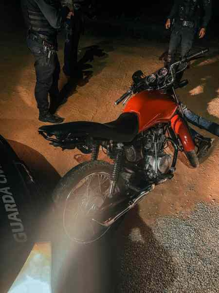 Motocicleta apreendida pela Guarda Municipal em Nova Serrana, com visíveis danos e sem placa, em cena de abordagem policial à noite.