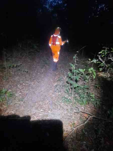 Um bombeiro vestindo um uniforme laranja, equipado com uma lanterna, caminha por um caminho estreito na floresta à noite, cercado por vegetação densa.