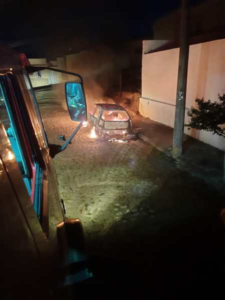 Um veículo Fiat Palio em chamas em uma rua estreita à noite, com fumaça saindo e fogo visível na parte traseira, observado de dentro de um caminhão de bombeiros.