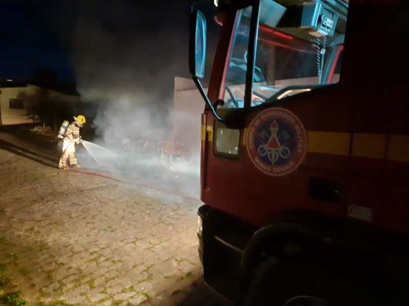 Bombeiro apagando incêndio em veículo, com fumaça ao redor e caminhão do Corpo de Bombeiros em primeiro plano.